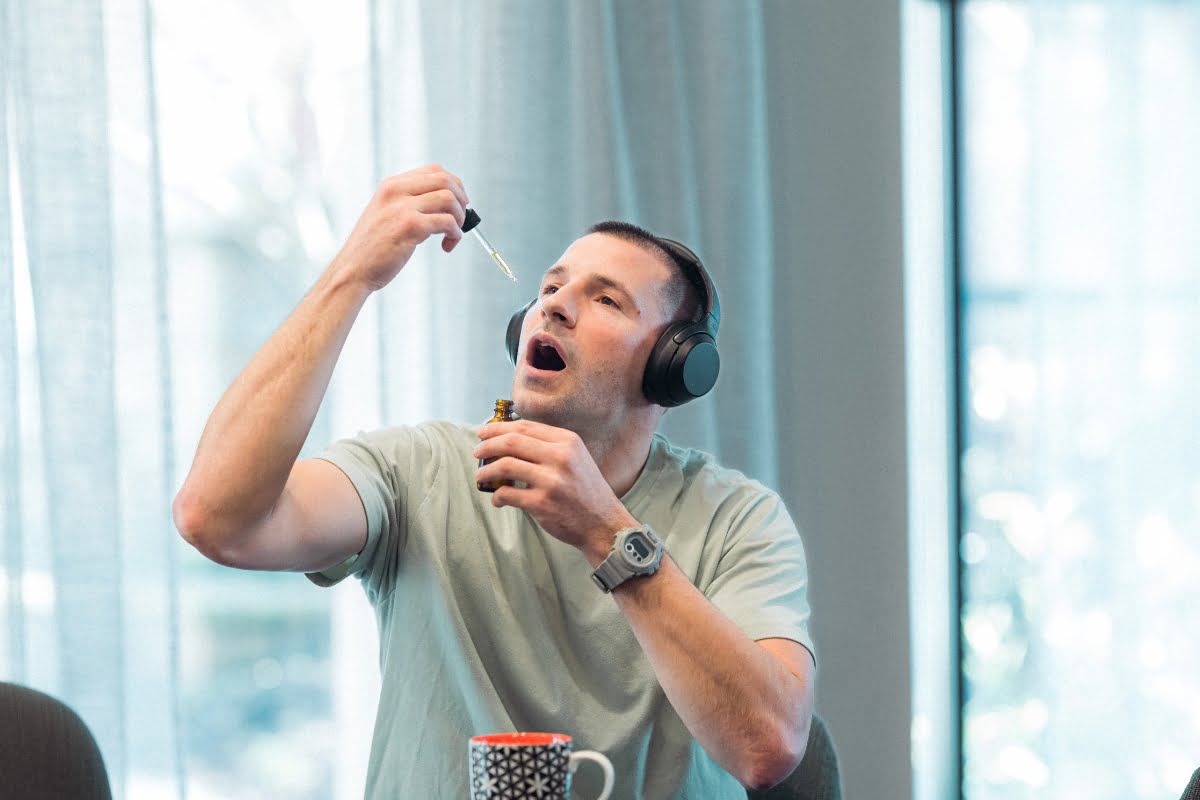 A person with headphones on is holding a CBD wellness dropper above their open mouth while sitting at a table with a mug beside them.