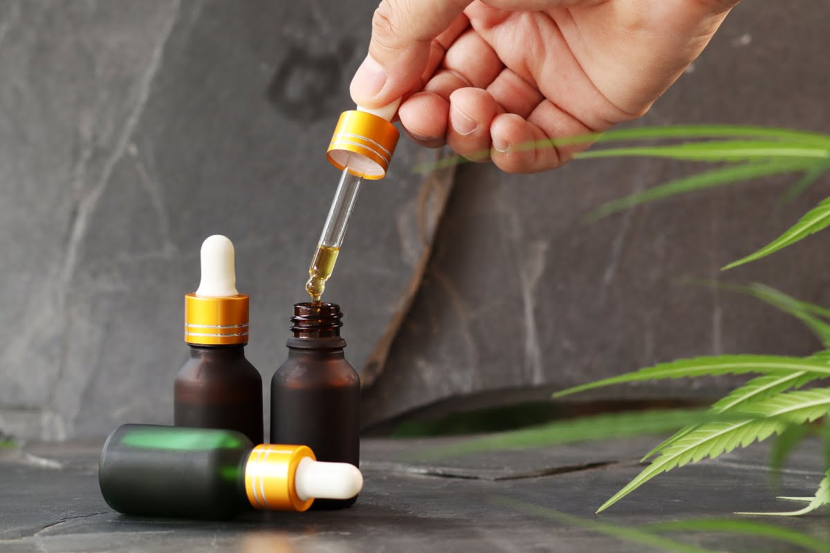 A hand holds a dropper above a small amber bottle, dispensing liquid. Two other similar bottles are nearby, and a green plant is partially visible in the foreground, evoking a serene cbd wellness scene.