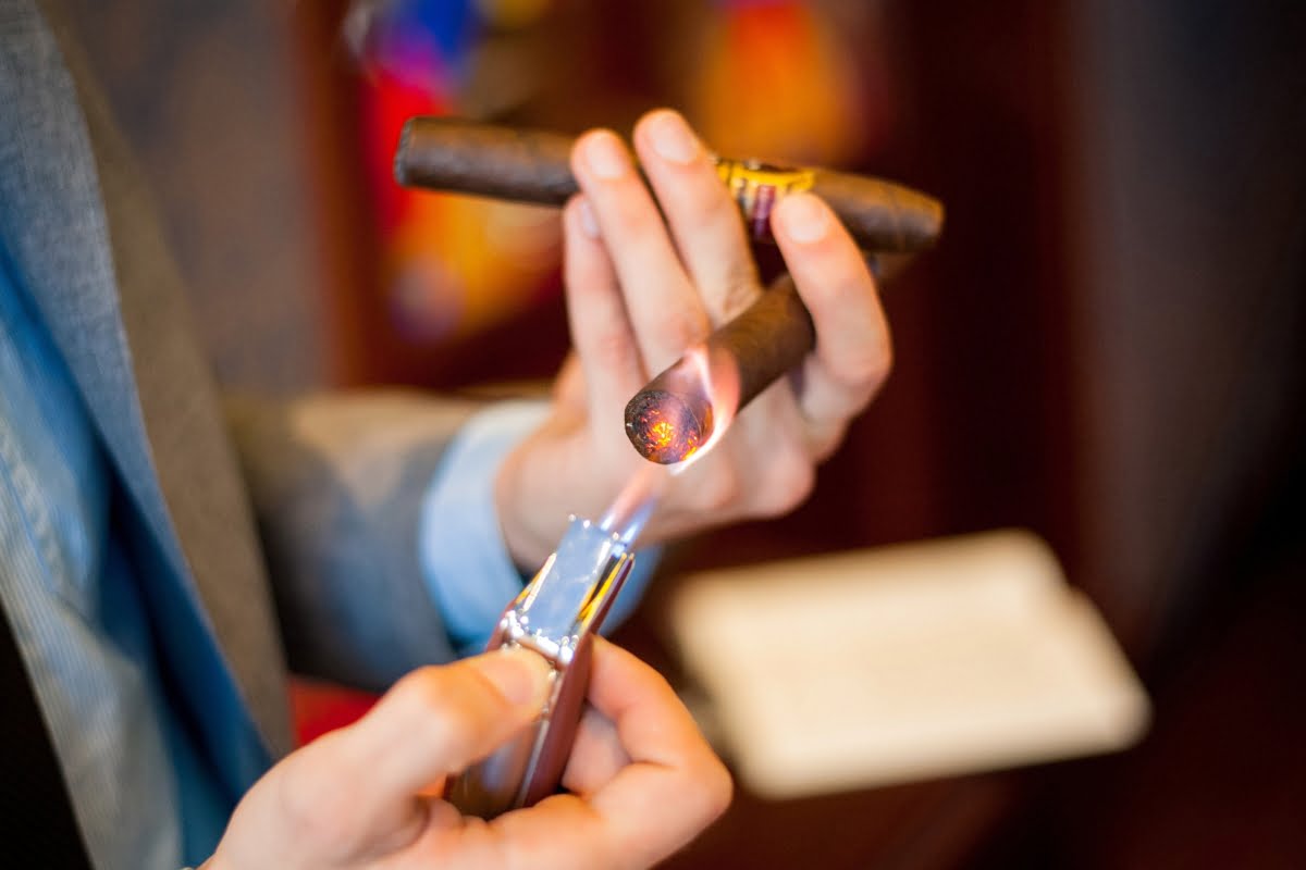 A smoking enthusiast in a suit lights a cigar with a lighter, holding another unlit cigar in the other hand.