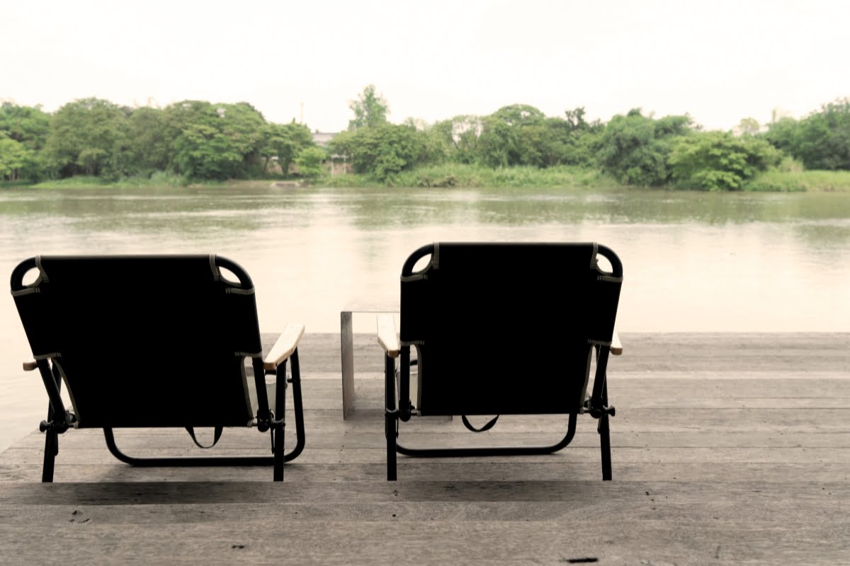 Two empty black lounge chairs on a wooden deck face a calm river with trees in the background, offering an ideal spot for a smoking enthusiast to unwind and enjoy the serene surroundings.