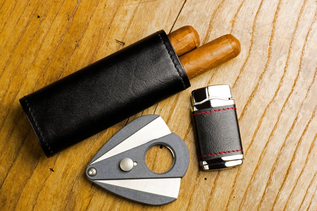 A black leather cigar case, ideal for the smoking enthusiast, lies on a wooden surface. Inside, it holds two cigars, a cigar cutter, and a metallic lighter with a matching black leather cover.