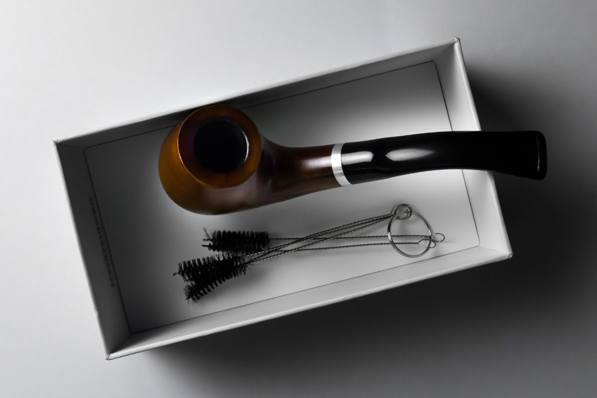 A smoking pipe and a set of pipe cleaners rest inside a white box placed on a gray surface, awaiting the discerning touch of a smoking enthusiast.