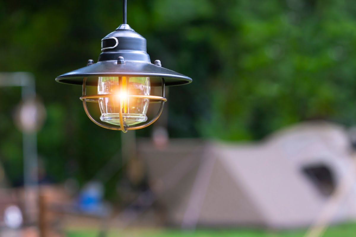 A hanging lantern is lit outdoors with a blurry background that includes a tent, perfect for any smoking enthusiast seeking a tranquil evening.