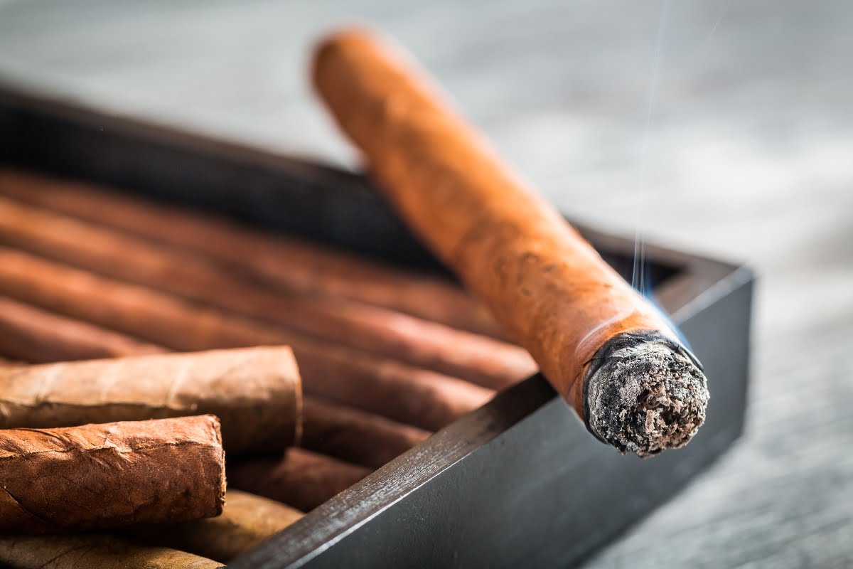 Close-up of a lit cigar resting on the edge of a wooden box filled with other cigars, perfect for any smoking enthusiast. Smoke is visible from the burning end.