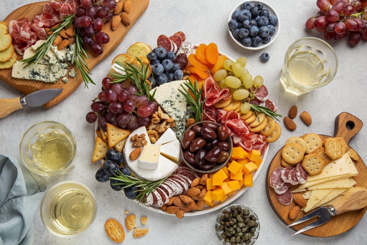 A charcuterie board with various cheeses, cured meats, grapes, blueberries, nuts, crackers, and cocktails. Surrounding it are additional plates of gourmet snacks like extra cheese, meats, bread, and glasses of white wine.