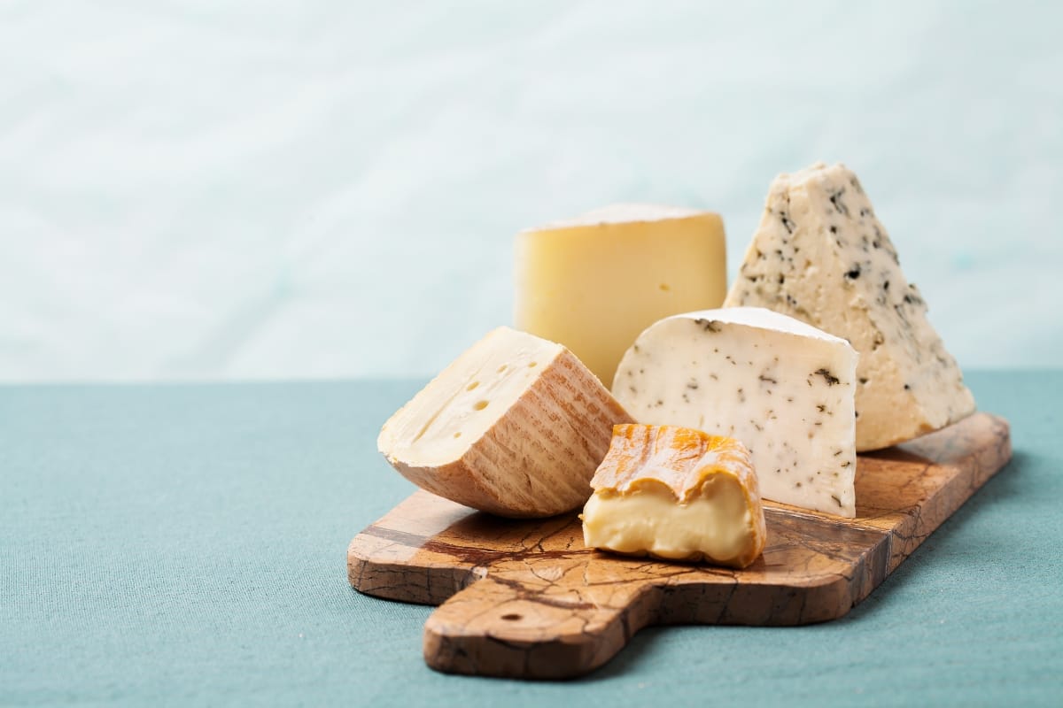 A variety of gourmet snacks, including cheeses, arranged on a wooden cutting board against a light blue background.