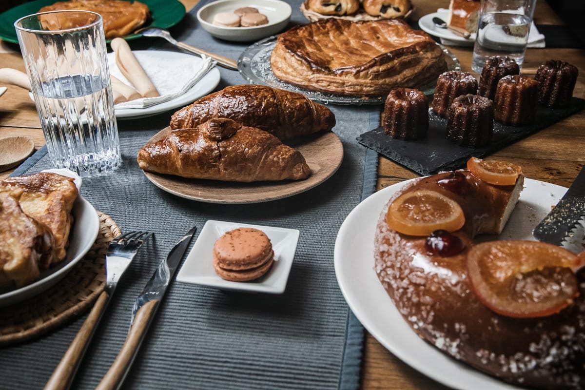 A table set with various gourmet snacks including croissants, a cake with candied fruit, macarons, and a glass of water.