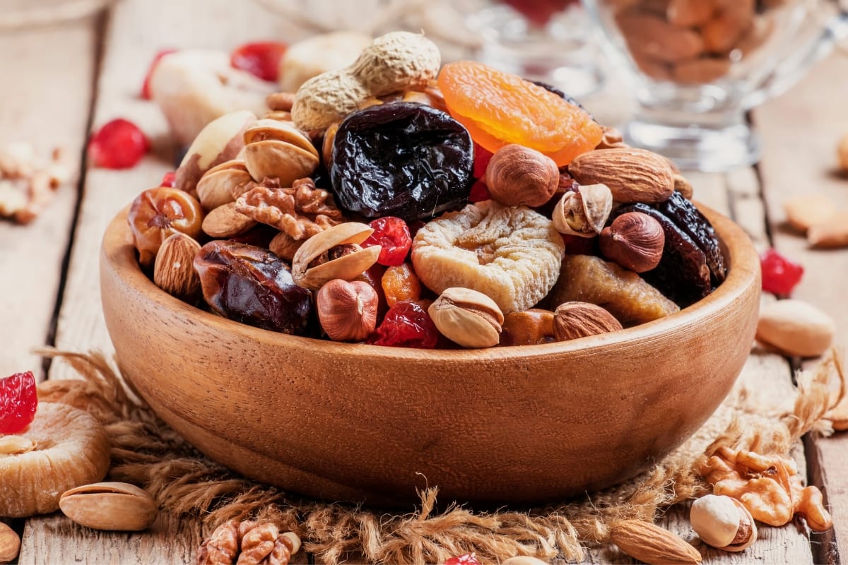 A wooden bowl filled with an assortment of gourmet snacks, featuring mixed nuts, dried fruits, and seeds on a rustic wooden surface.