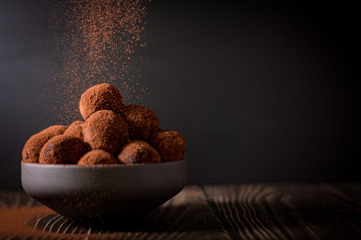 A bowl of round chocolate truffles, a delightful addition to any collection of gourmet snacks, is topped with cocoa powder being sprinkled from above against a dark background on a wooden surface.