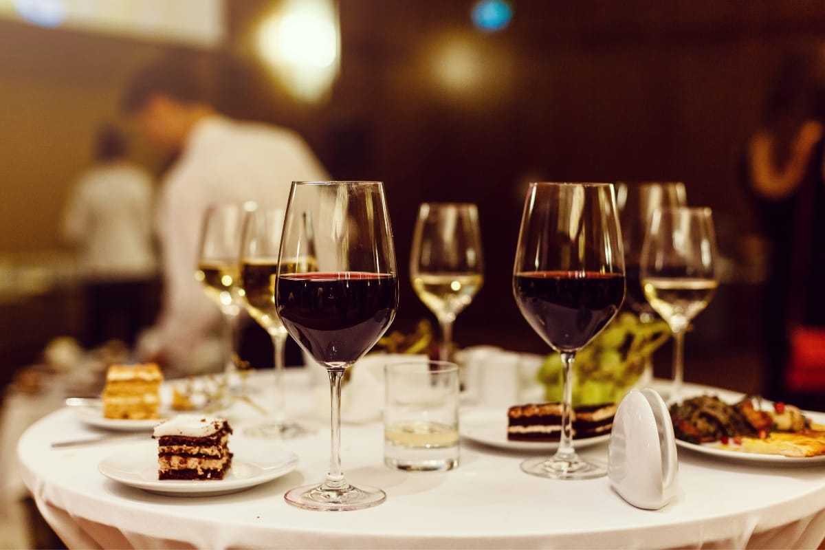 A close-up of a table with several glasses of wine, pieces of cake, and plates of gourmet snacks. People can be seen in the background.