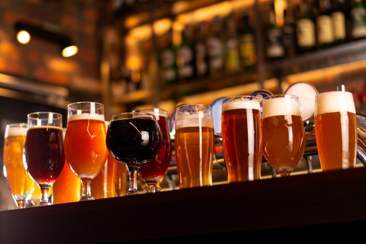 A row of various beer glasses filled with different types of beer is displayed on a bar counter, accompanied by gourmet snacks, with a blurred background of bottles and taps.