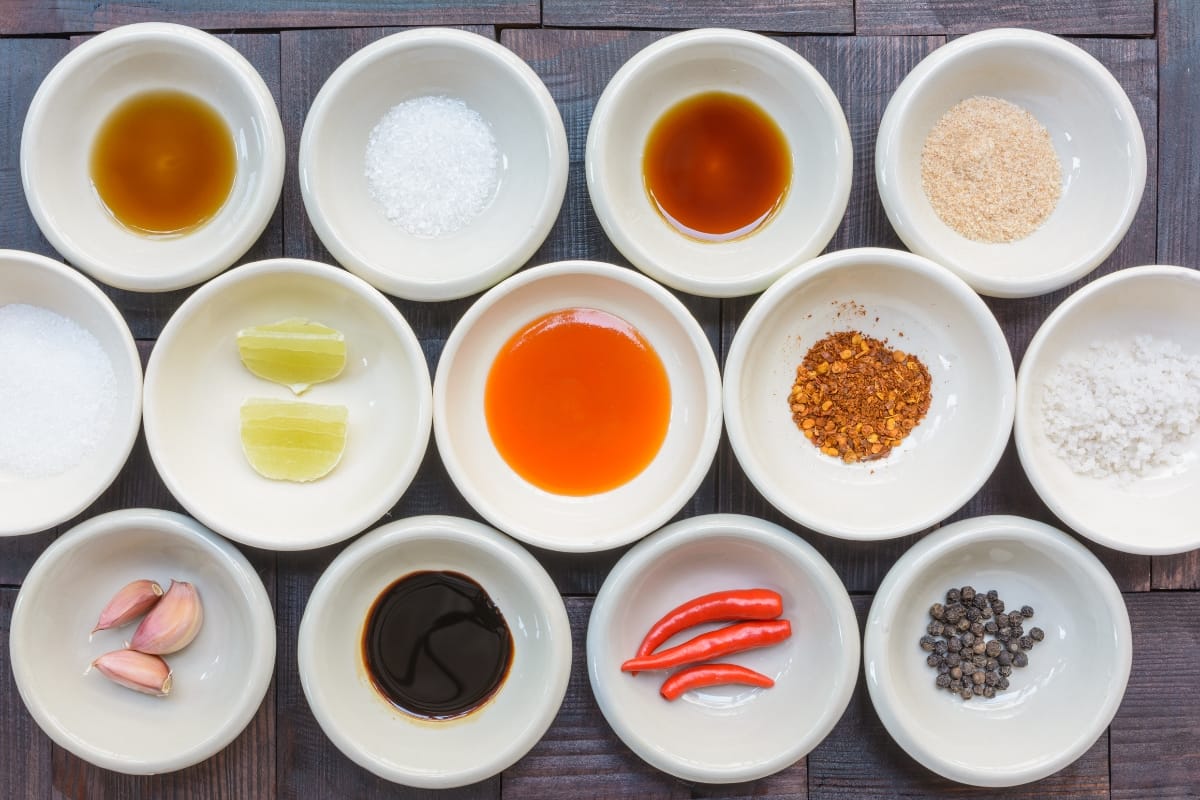 An assortment of various condiments and spices in small bowls arranged on a wooden surface, including olive oil, salt, soy sauce, lime, chili flakes, garlic cloves, whole chili peppers, and peppercorns—perfect accompaniments for your gourmet snacks.