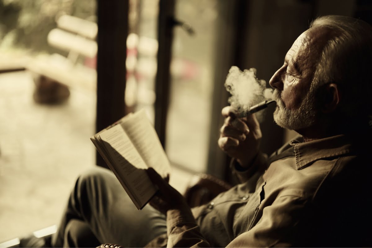 Elderly man with grey hair and beard reading a book during a relaxed smoking session indoors.