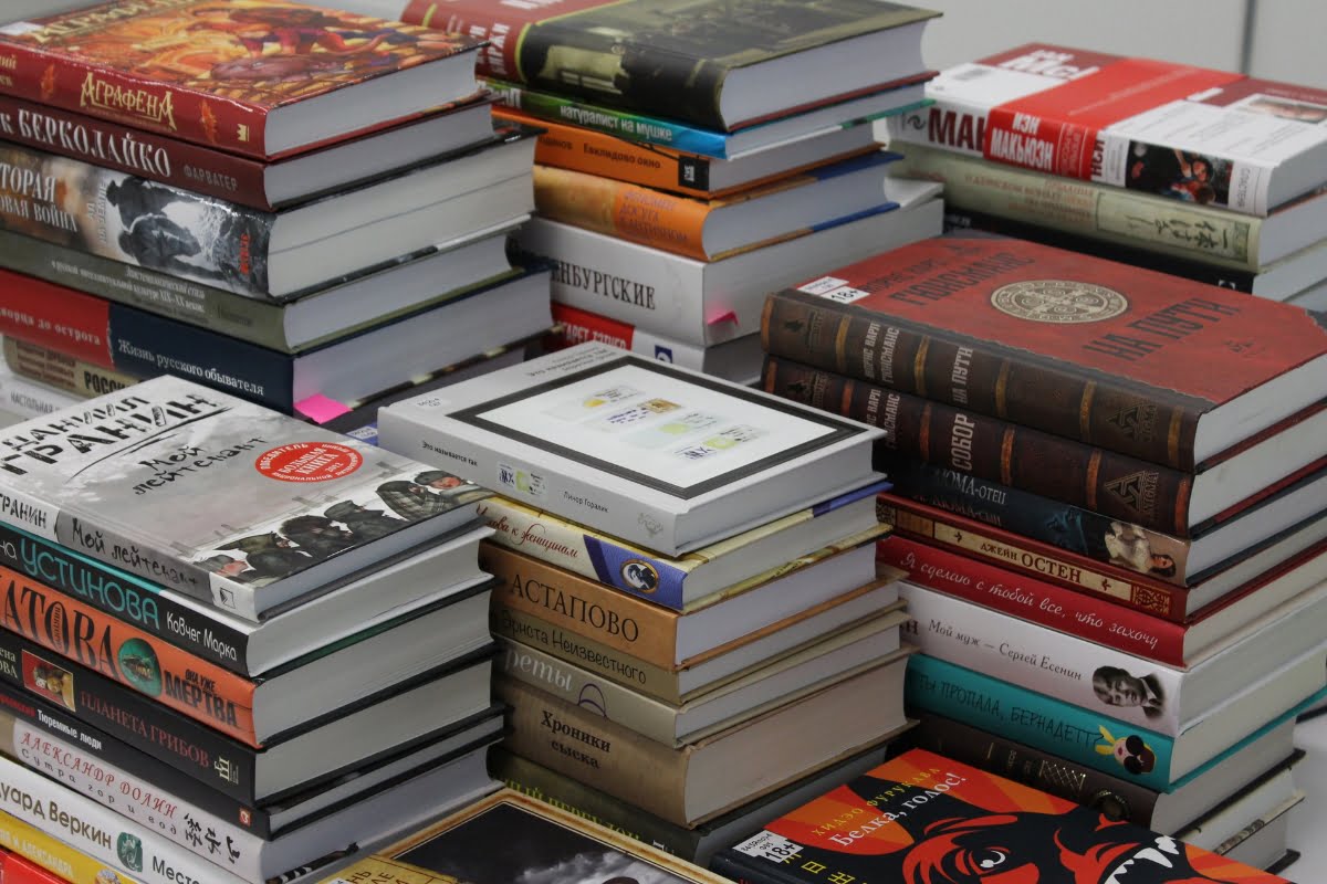 A large collection of stacked books in various languages, with a single e-reader lying on top of one of the stacks, creates the perfect ambiance for a cozy smoking session.