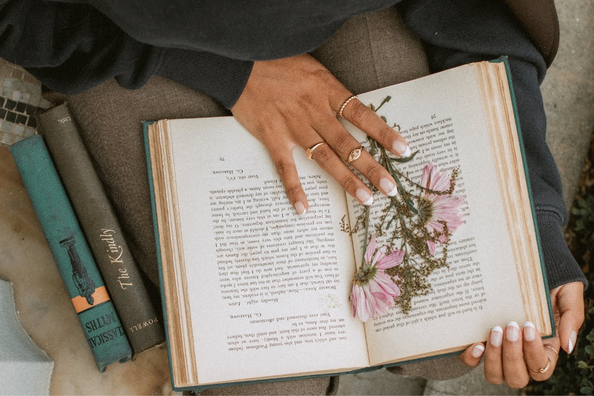 A person with multiple rings holds an open book with pressed pink flowers inside, as the faint scent from a recent smoking session lingers. There are additional books lying partially visible next to them.