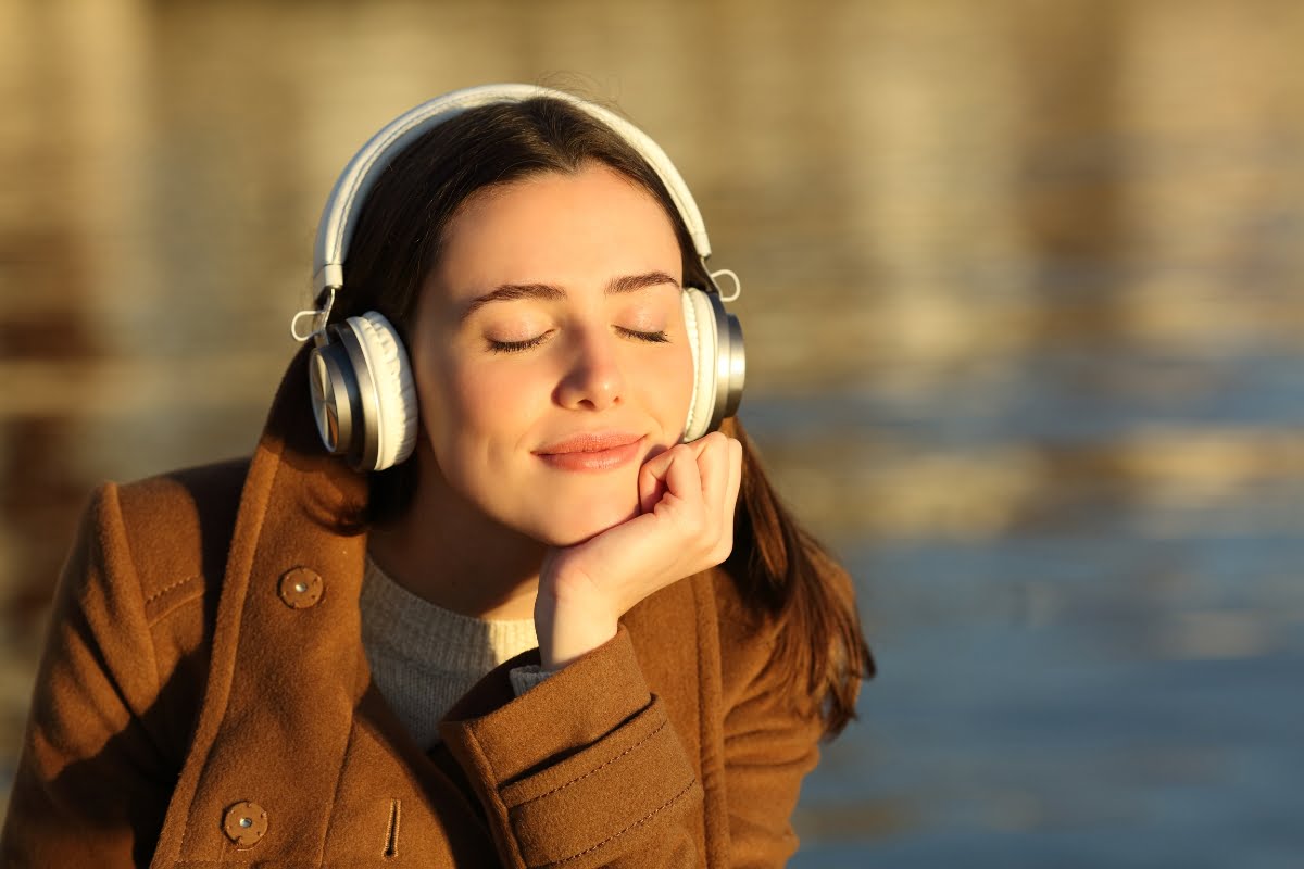 A woman wearing a brown coat and large headphones smiles with her eyes closed, resting her head on her hand, as if reminiscing about a peaceful smoking session, in front of a blurred water background.