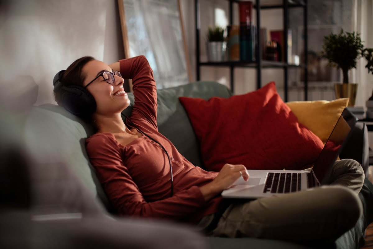 A person wearing glasses and headphones lounges on a couch with one hand on a laptop, surrounded by colorful cushions, smiling and appearing relaxed after a smoking session.