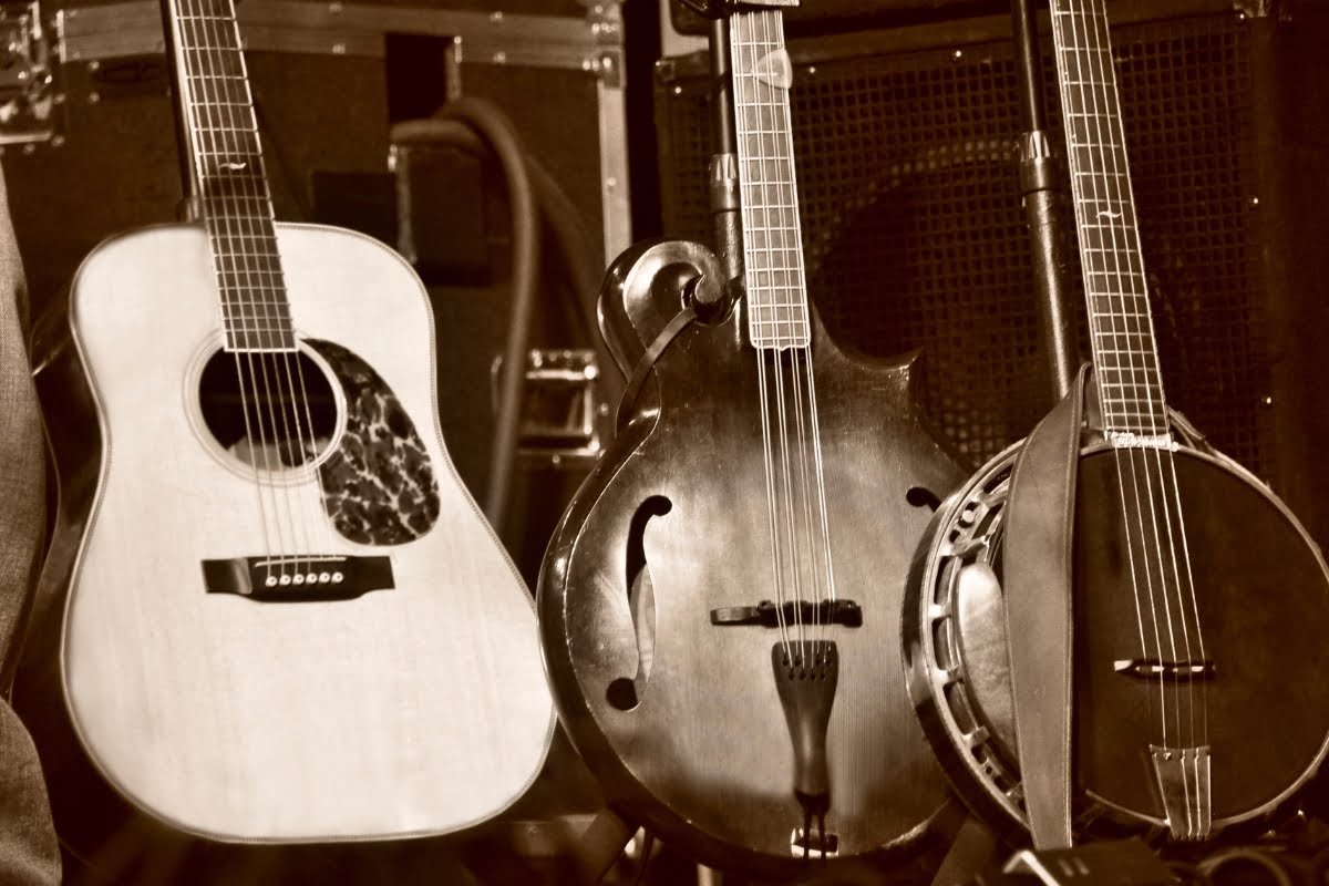 A sepia-toned image showing three string instruments: an acoustic guitar, a mandolin, and a banjo, standing upright against a backdrop of audio equipment, evokes the vibe of an old-time smoking session.