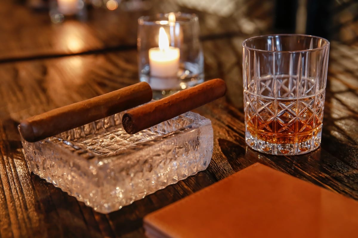 A glass of artisanal whiskey and two cigars on a crystal ashtray rest on a wooden table next to a burning candle.