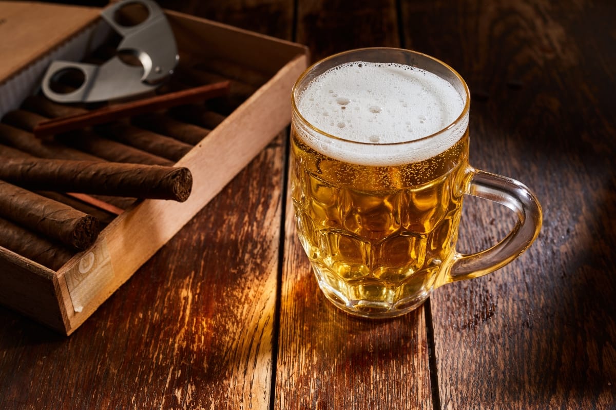 An artisanal beverage, the glass mug of beer rests on a wooden table, accompanied by an open box of cigars and a cigar cutter.