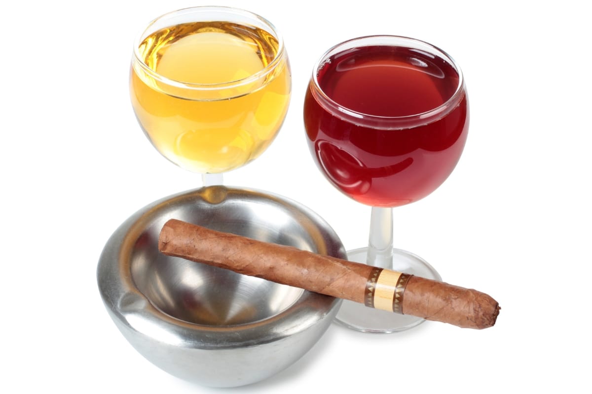 Two glasses of artisanal wine, one white and one red, sit elegantly next to a cigar resting in a metal ashtray on a pristine white background.