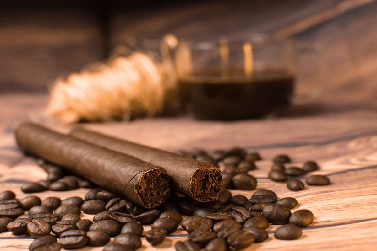 Two cigars rest on a wooden surface, surrounded by coffee beans, while a glass cup of artisanal coffee graces the background.