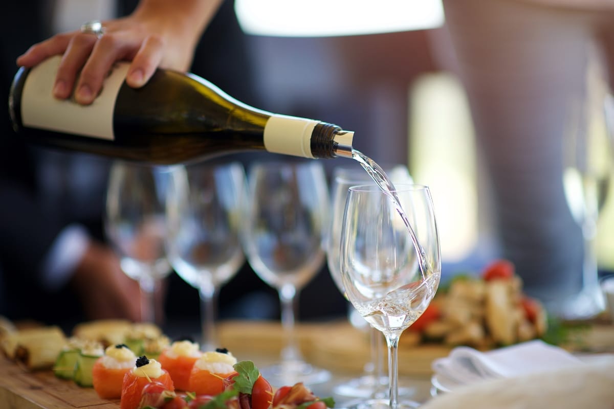 A person pours artisanal white wine into a glass on a table set with assorted appetizers.