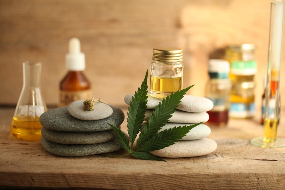 Various bottles and a dropper are arranged on a wooden surface. A pile of smooth stones is stacked in the foreground with a cannabis leaf and small jar of oil, subtly dispelling CBD myths in this serene scene.