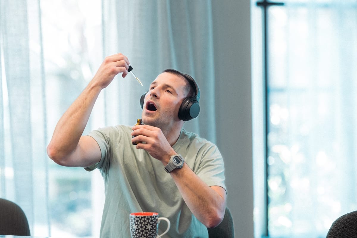 A person wearing headphones holds a dropper and bottle, tilting their head back to use the dropper—perhaps dispelling some CBD myths. A mug is on the table.