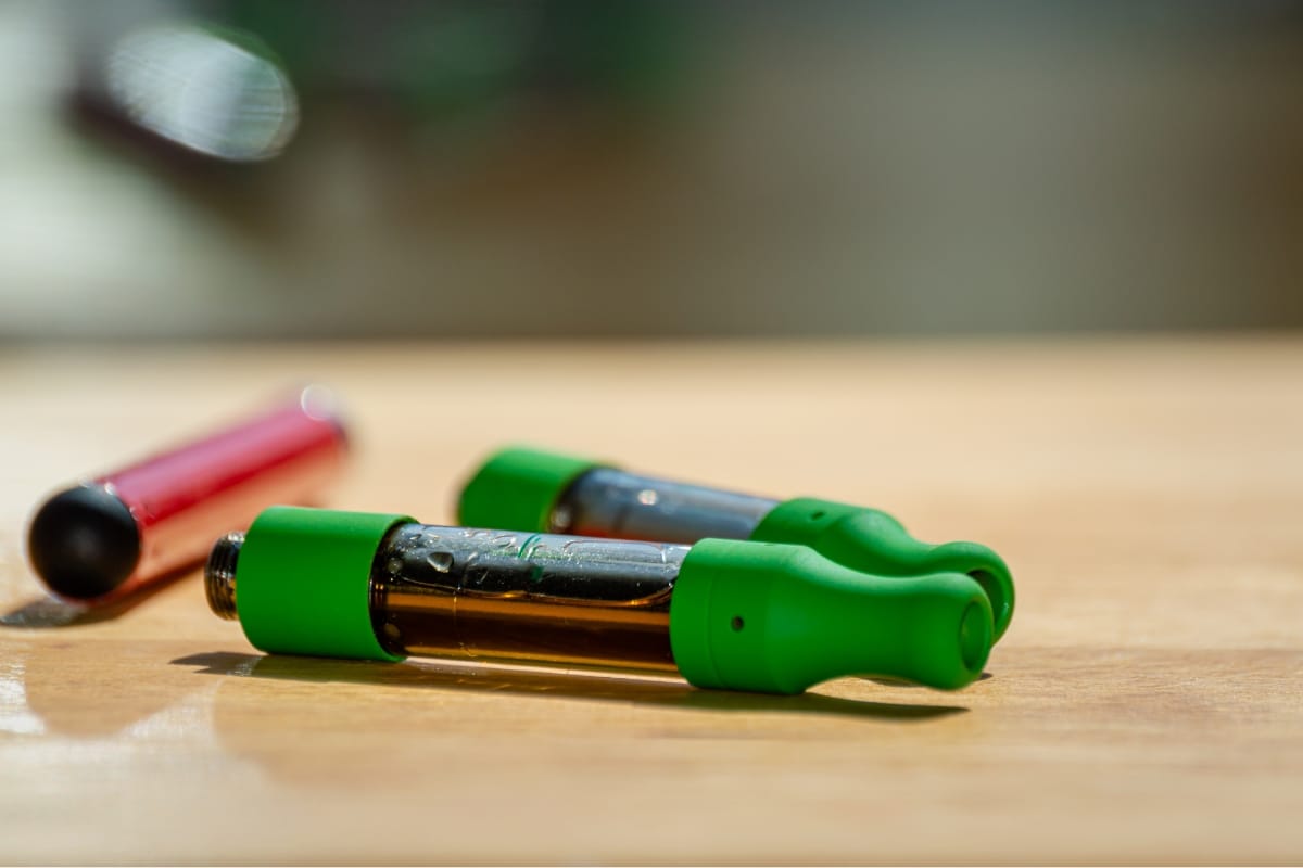 Two CBD vape pens with green mouthpieces rest on a wooden surface, next to a red cylindrical object.