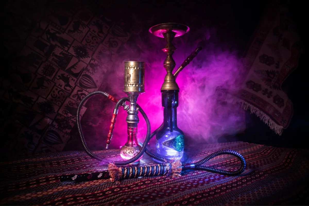 Two hookahs sit elegantly on a patterned surface, enveloped by swirling purple and pink smoke. The decorative backdrop accentuates the age-old allure of hookahs, inviting whispers of charm in a world now curious about hookah vs vape debates.