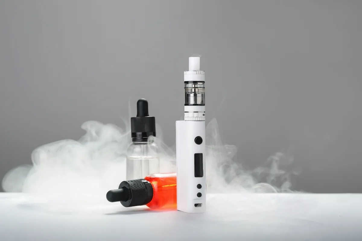 White vape device surrounded by two dropper bottles, one with red liquid and the other clear; smoke drifts around them against a gray background, offering a serene glimpse into the hookah vs vape debate.