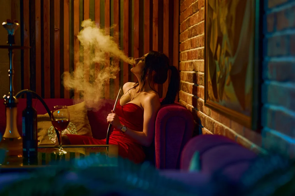 In a dimly lit room, a woman in a red dress sits on a purple couch, exhaling smoke from her hookah while pondering the hookah vs vape debate. A glass of red wine rests on the table, capturing the evening's relaxed allure.